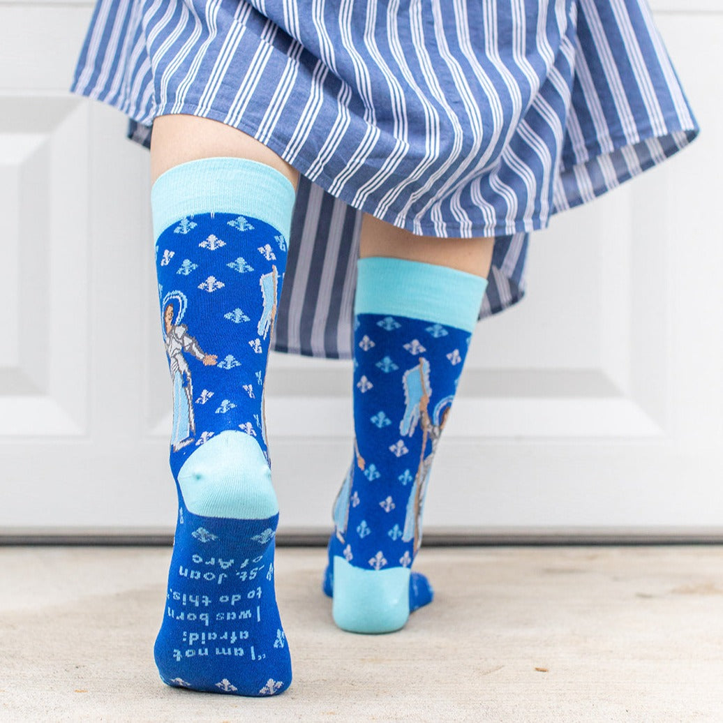 a woman wearing blue dress socks with St. Joan of Arc on them with a fleur de lis pattern on them showing the bottom of the sock