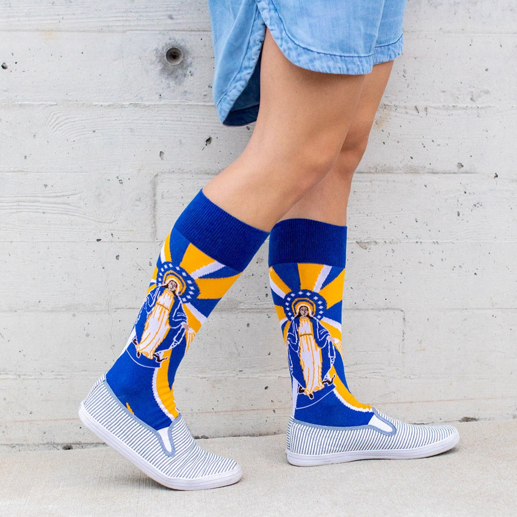 woman wearing Our Lady of Grace socks with Mary, yellow and blue rays and a serpant on the bottom of the foot with white shoes on
