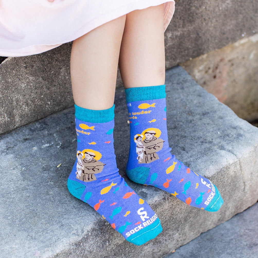 little girl sitting on steps wearing blue socks featuring St. Anthony holding the infant Jesus with several fish and the words "Actions speak louder than words" along the calf