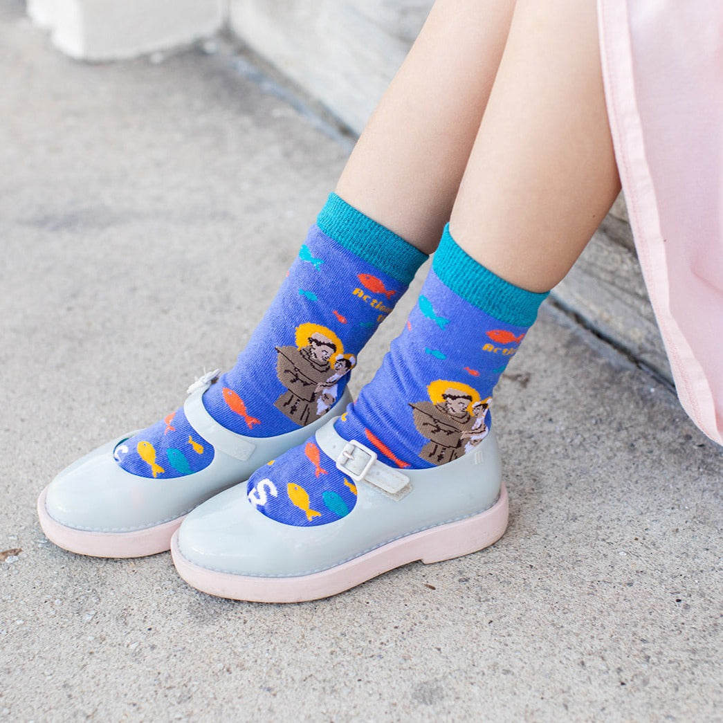 little girl wearing gray dress shoes and blue socks featuring St. Anthony holding the infant Jesus with several fish and the words "Actions speak louder than words" along the calf