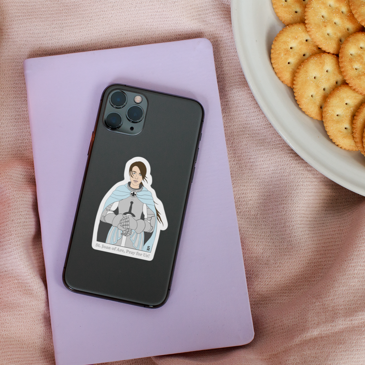 Sticker with St. Joan of Arc in full armor with a blue cape that says "St. Joan of Arc, pray for us" along the bottom on a phone