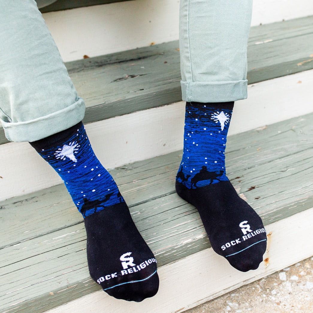 man sitting on steps wearing Black and blue socks featuring silhouettes of the 3 wisemen on camels following a bright white star of bethlehem