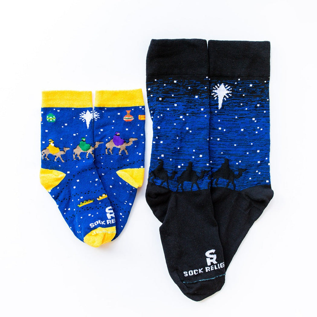 Black and blue socks featuring silhouettes of the 3 wisemen on camels following a bright white star of bethlehem next to the kids version that features more bright colors like yellow, green and purple