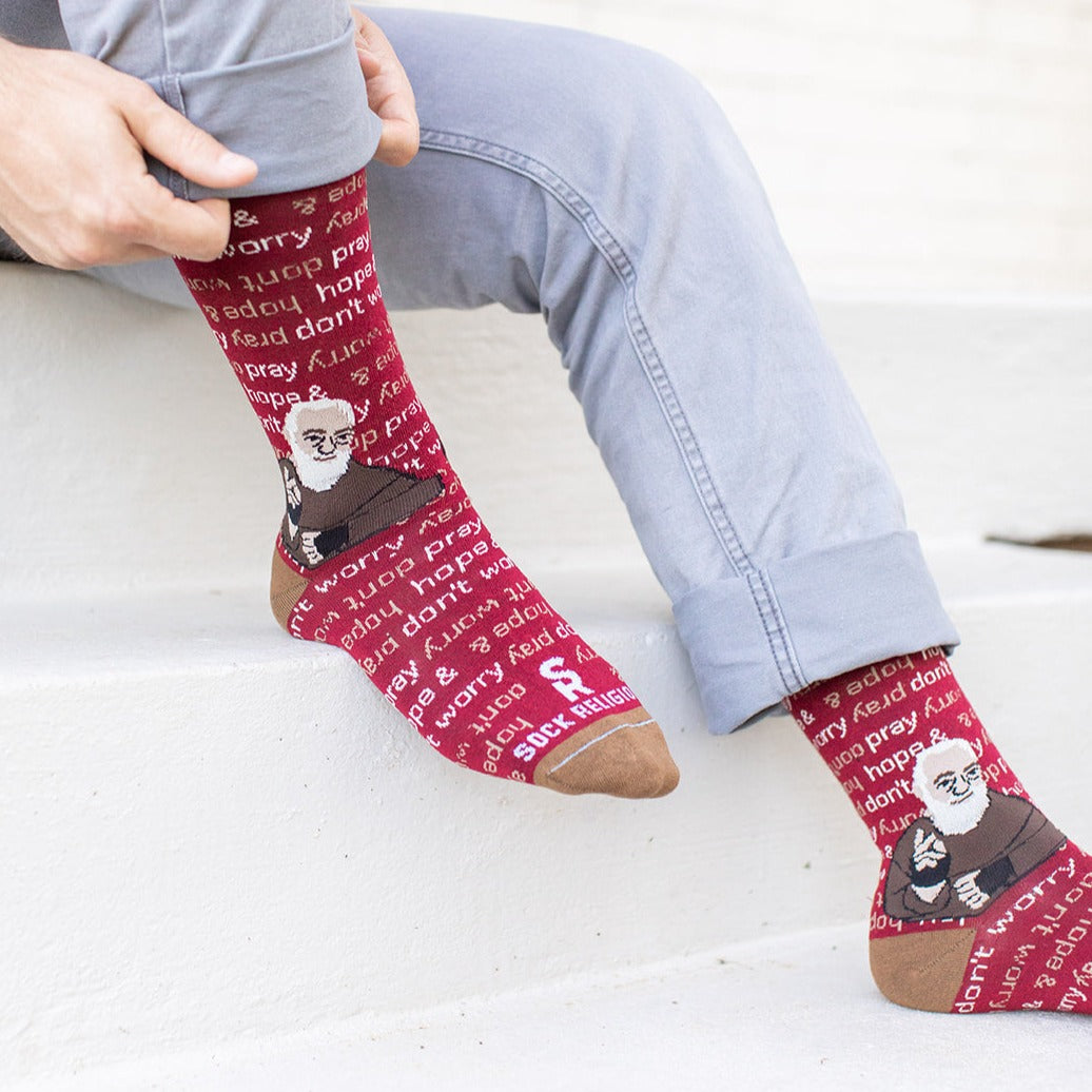 man sitting on steps wearing maroon and light brown socks that repeat 'pray, hope, and don't worry' in the background and feature St. Padre Pio