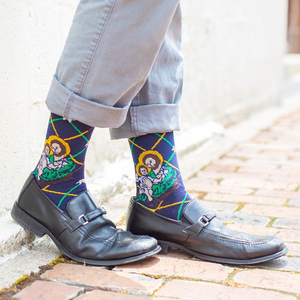 man in black dress shoes wearing Navy blue St. Joseph adult XL socks with Joseph holding baby Jesus. Yellow and green argyle lines