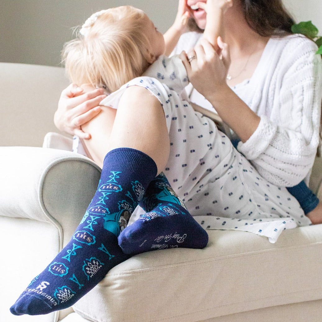 a woman sitting on a couch holding a baby wearing a pair of blue socks with a picture of St. Monica