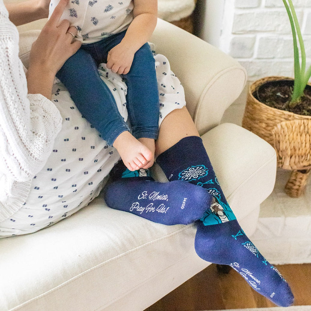a woman sitting on a couch holding a small child wearing a pair of blue socks with a picture of St. Monica
