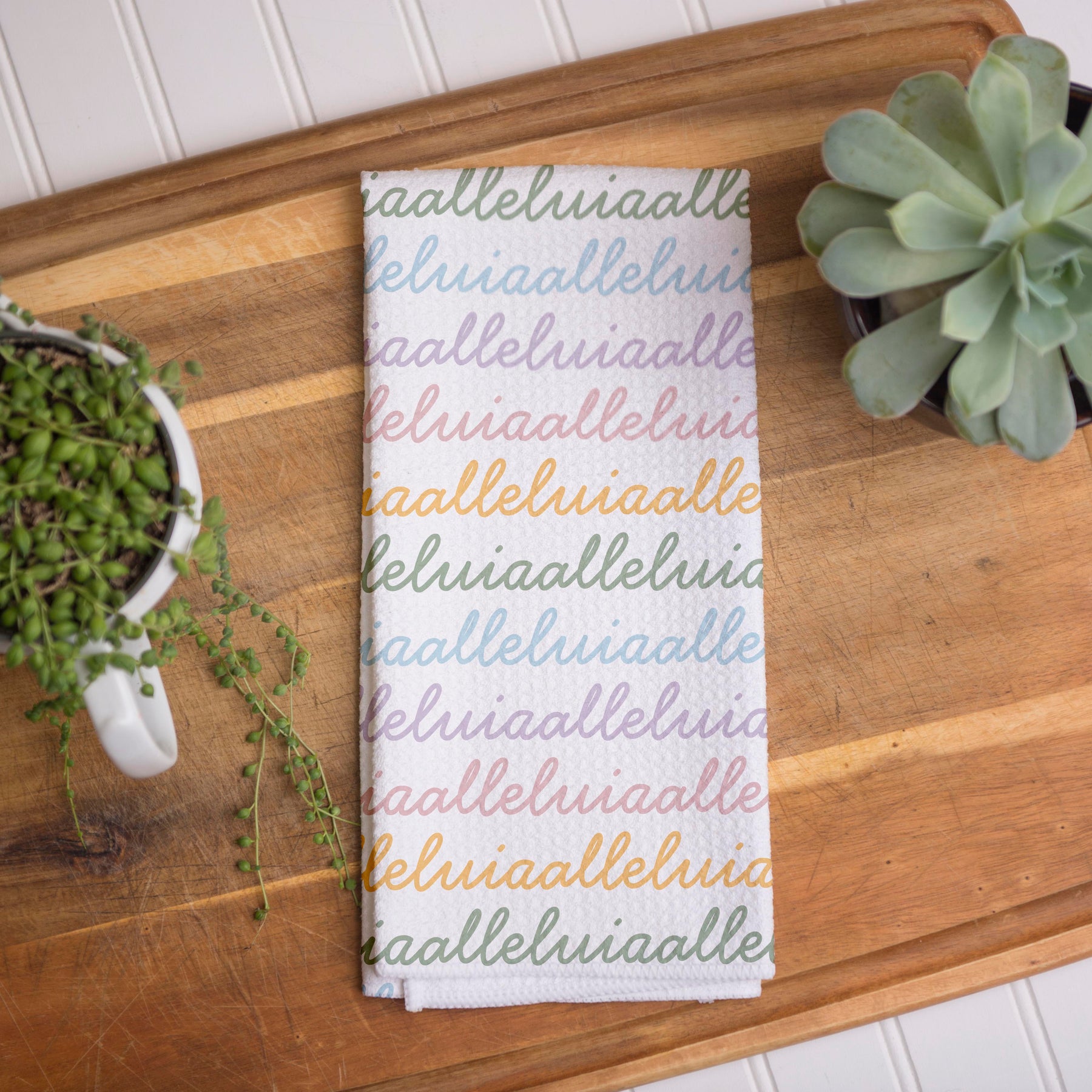 White colored, waffle textured dish towel folded in half with the design that says alleluia, alleluia repeatedly in cursive writing, in colors pastel pink, pastel yellow, pastel blue and pastel green.