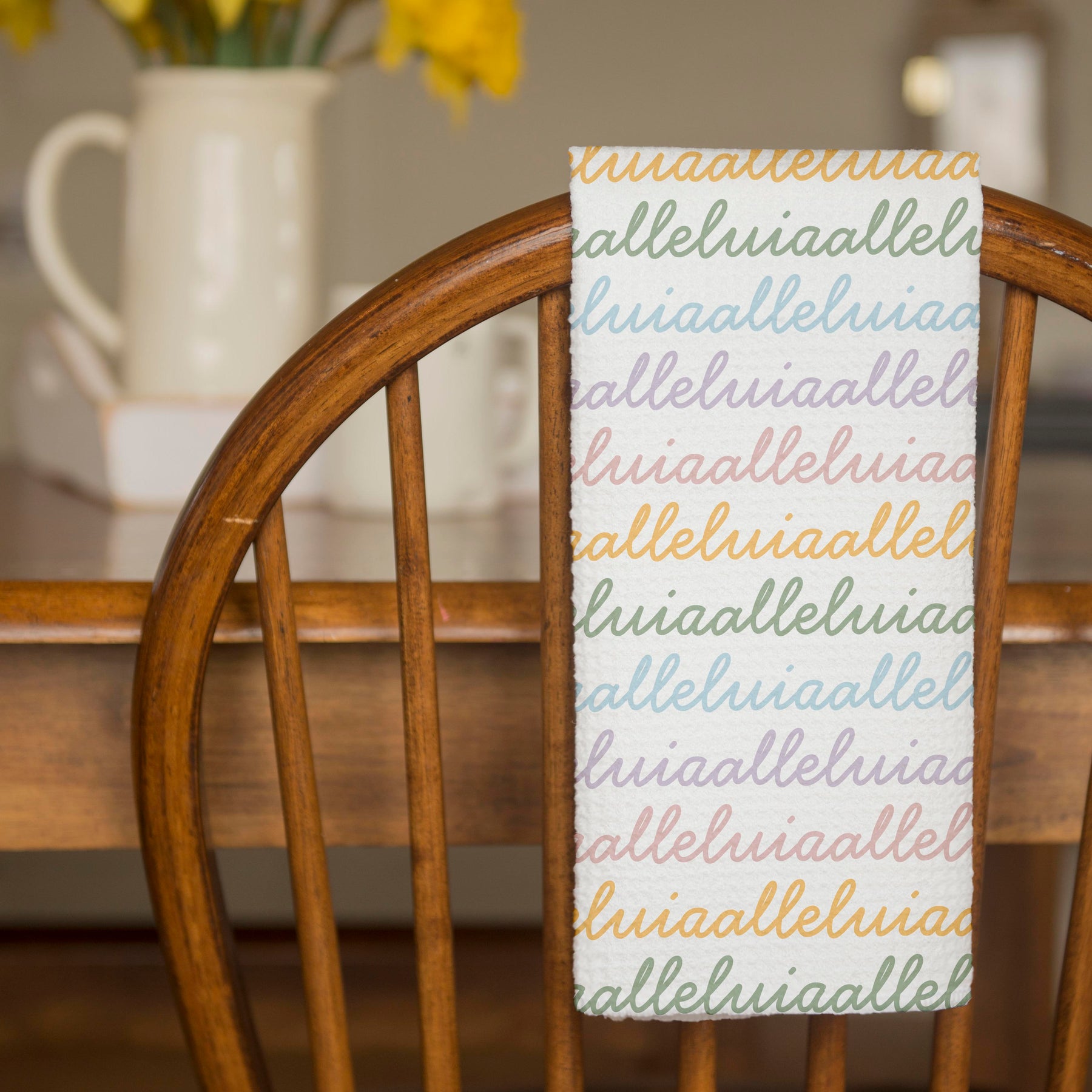 White colored, waffle textured dish towel folded in half, hanging on wooden a chair, with the design that says alleluia, alleluia repeatedly in cursive writing, in colors pastel pink, pastel yellow, pastel blue and pastel green.