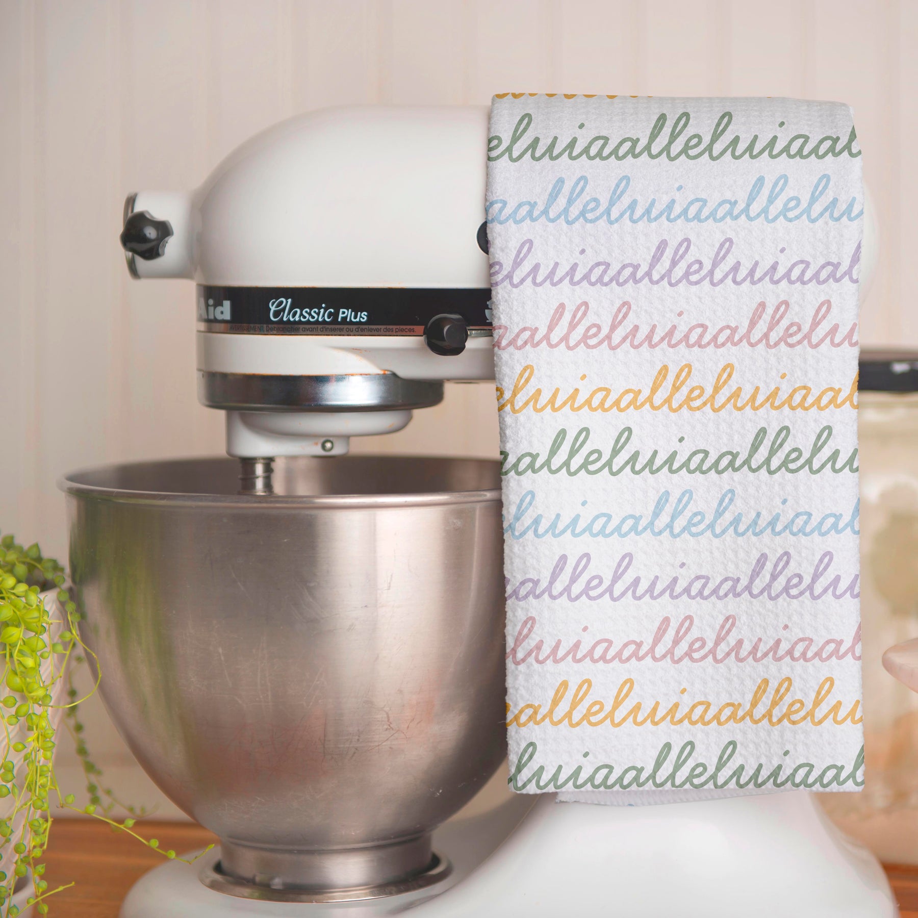 White colored, waffle textured dish towel folded in half, hanging on a white kitchen mixer, with the design that says alleluia, alleluia repeatedly in cursive writing, in colors pastel pink, pastel yellow, pastel blue and pastel green.