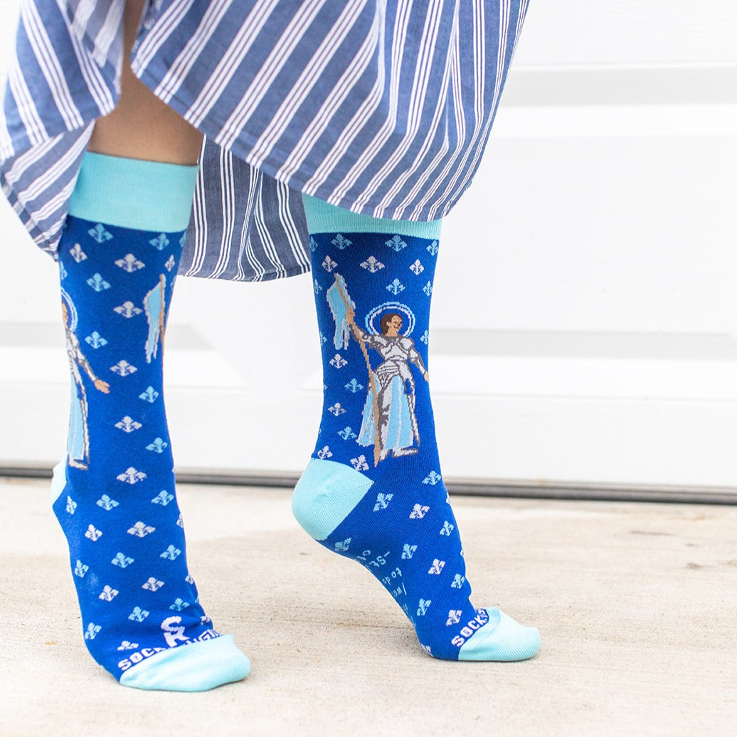 a woman standing wearing blue dress socks with St. Joan of Arc on them with a fleur de lis pattern on them 