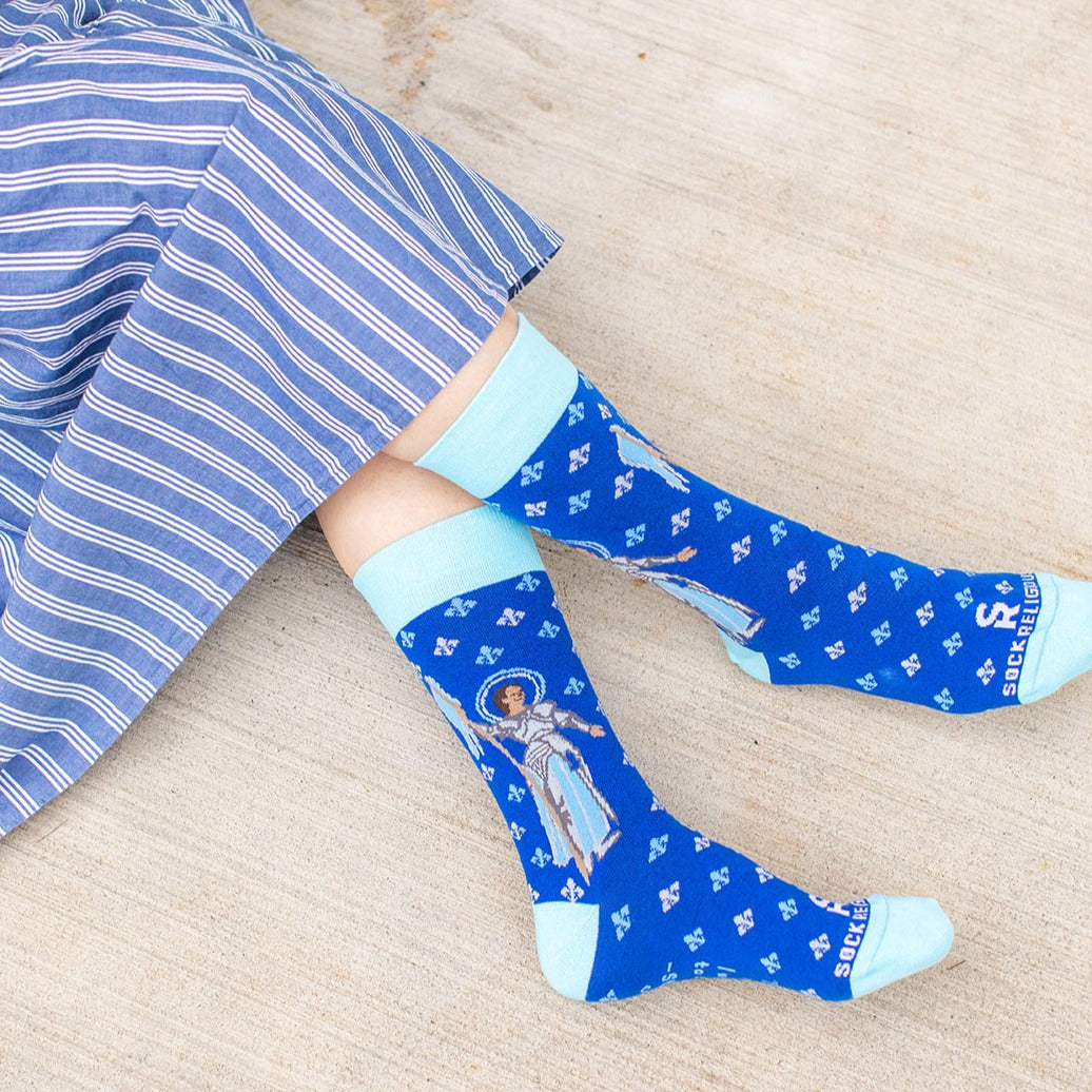 a woman sitting wearing blue dress socks with St. Joan of Arc on them with a fleur de lis pattern on them 