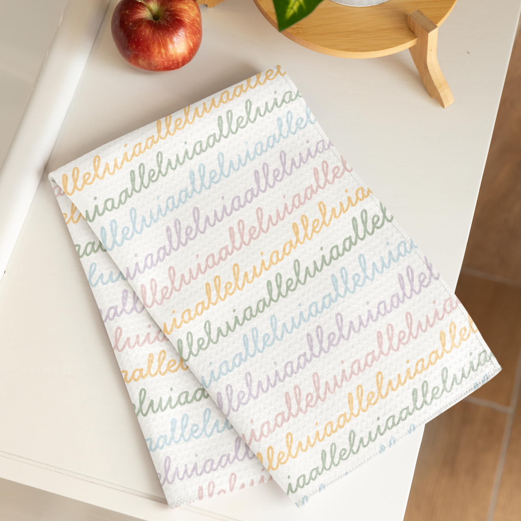 White colored, waffle textured dish towel folded in half with the design that says alleluia, alleluia repeatedly in cursive writing, in colors pastel pink, pastel yellow, pastel blue and pastel green.