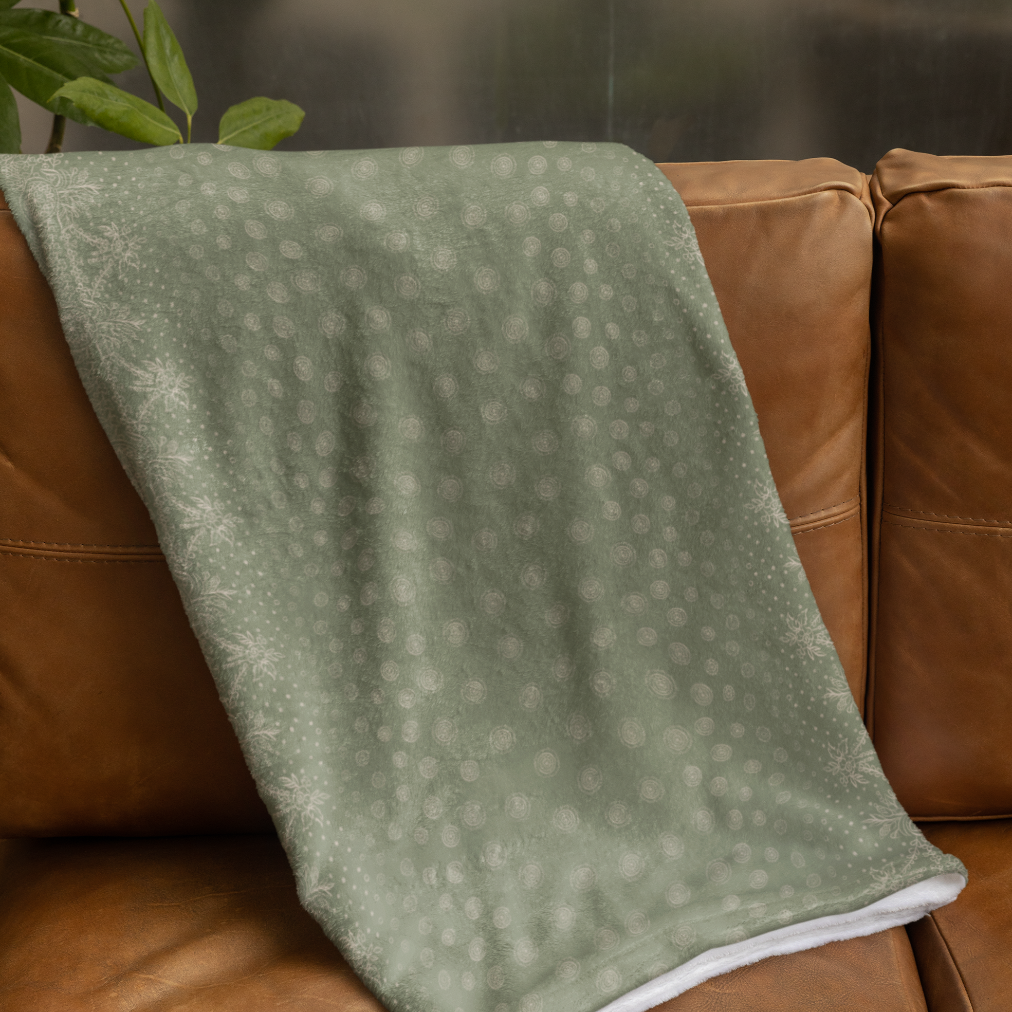 A green and gold blanket with a white backside, showcasing a gold design of craft symbols by Louis and Zélie placed on a brown leather couch.